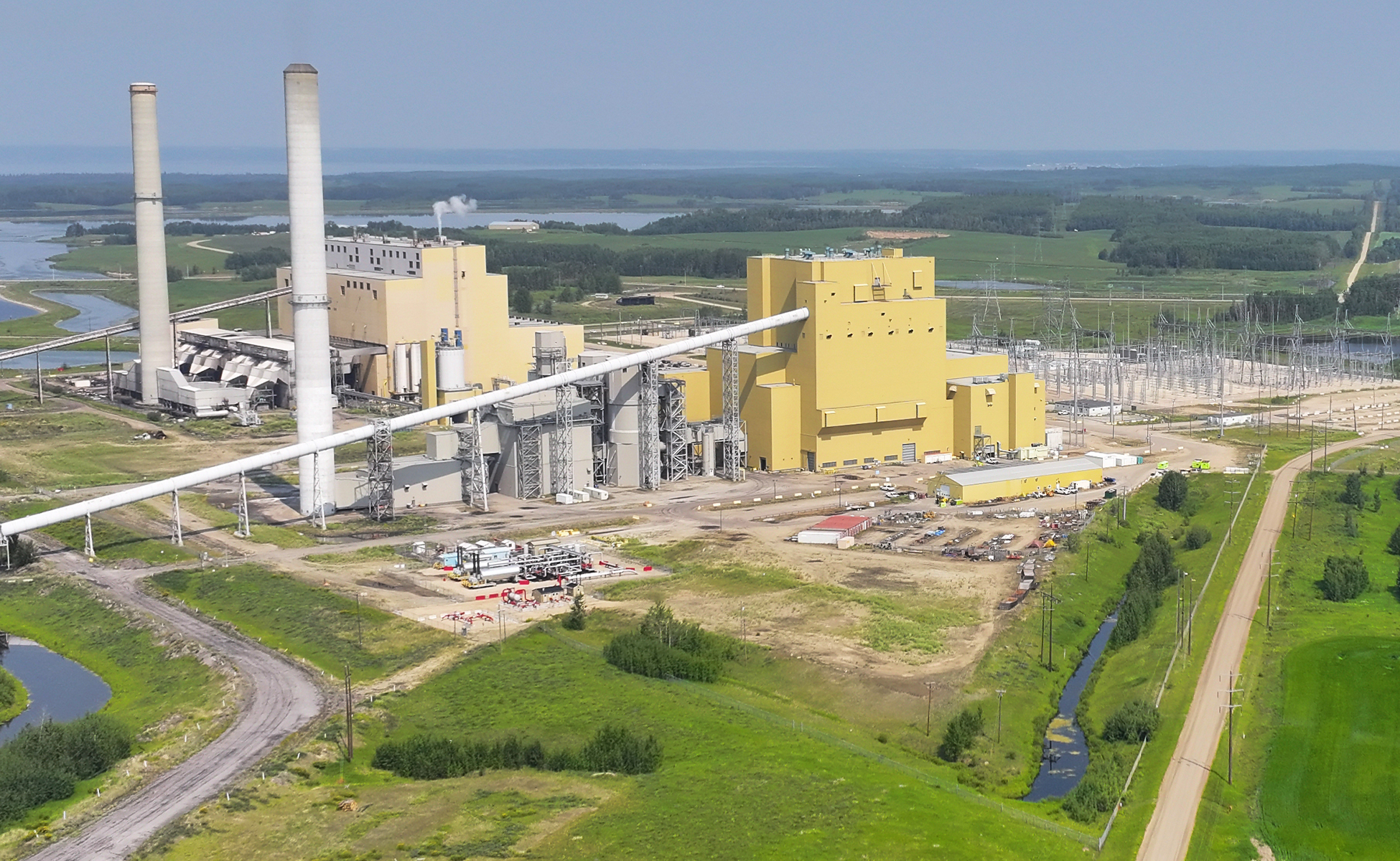 Aerial view of a large industrial power plant complex with two tall smokestacks and multiple yellow buildings, surrounded by electrical infrastructure, green fields, a winding road, and a nearby waterway under a clear sky.