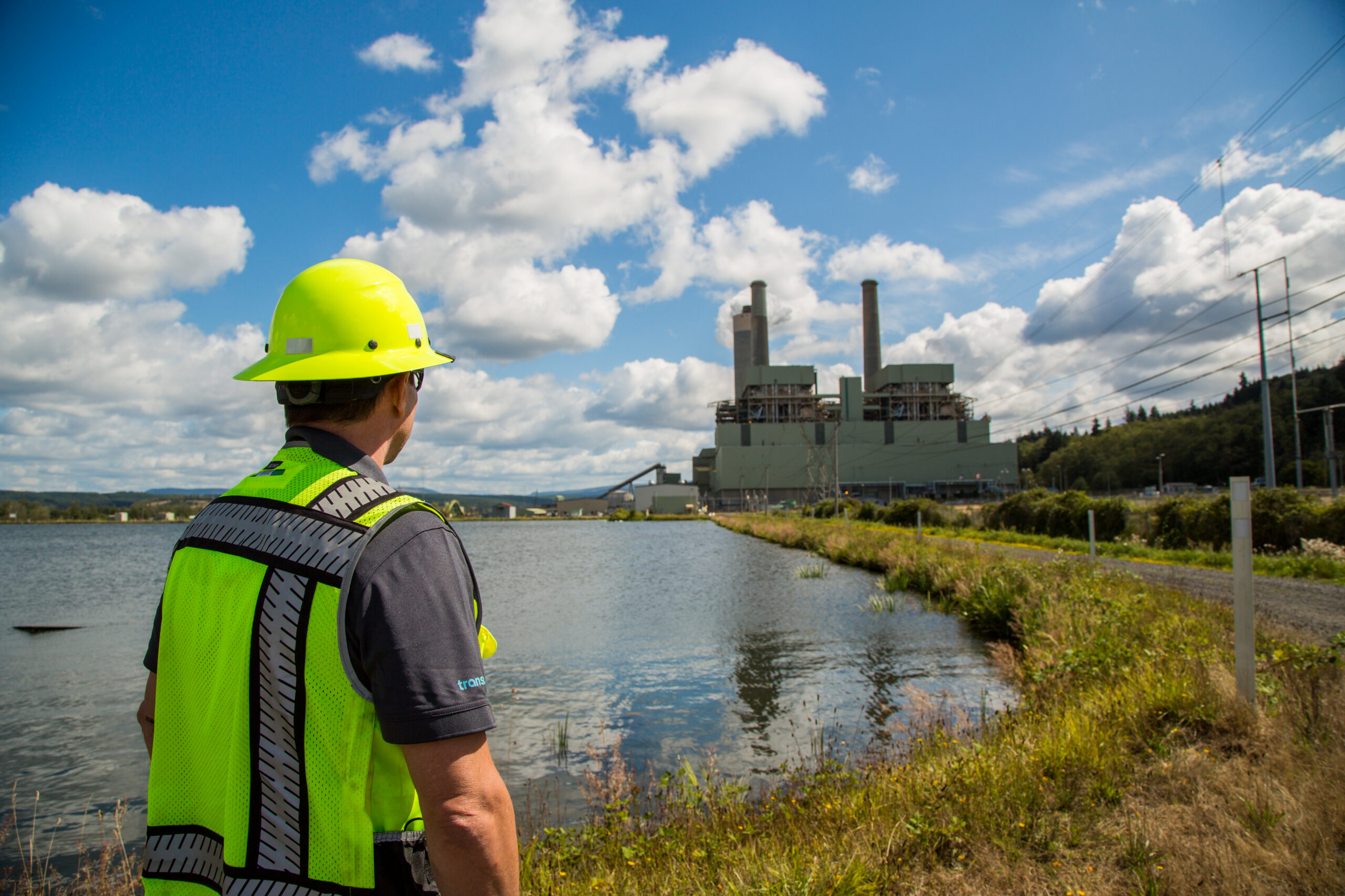 man in hard hat and safety vest looking out at a pond and coal plant facility in the distance. The sky is blue and cloudy.